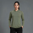 A woman modeling an olive green long-sleeve athletic hoodie with white piping details and a small logo on the left chest, paired with black pants, against a solid gray studio background.